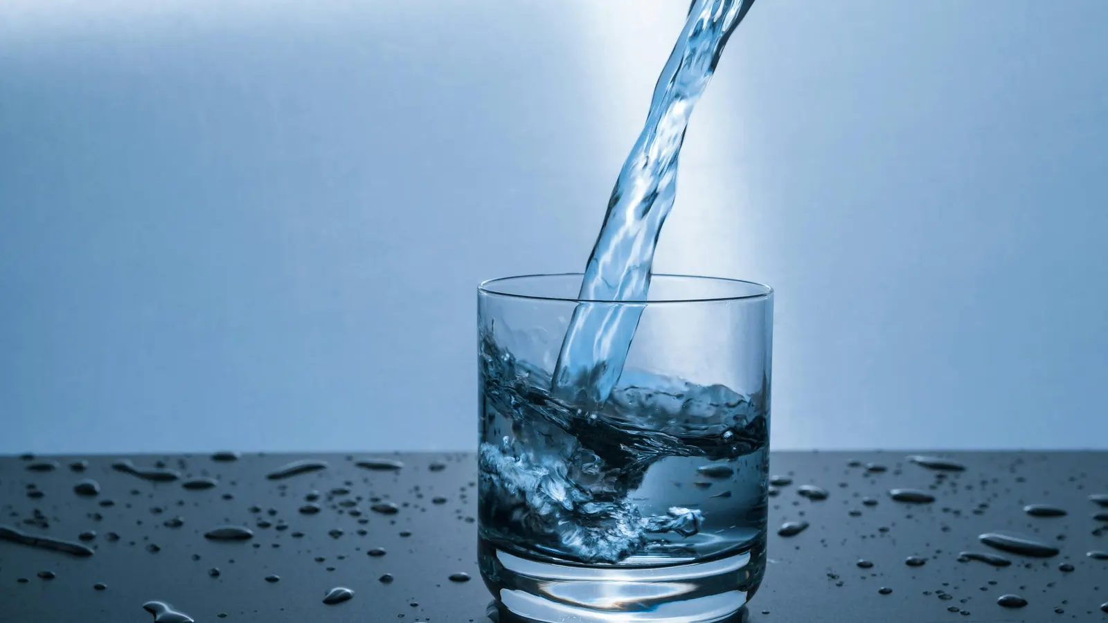 Water pouring into a clear glass tumbler on a dark countertop scattered with droplets