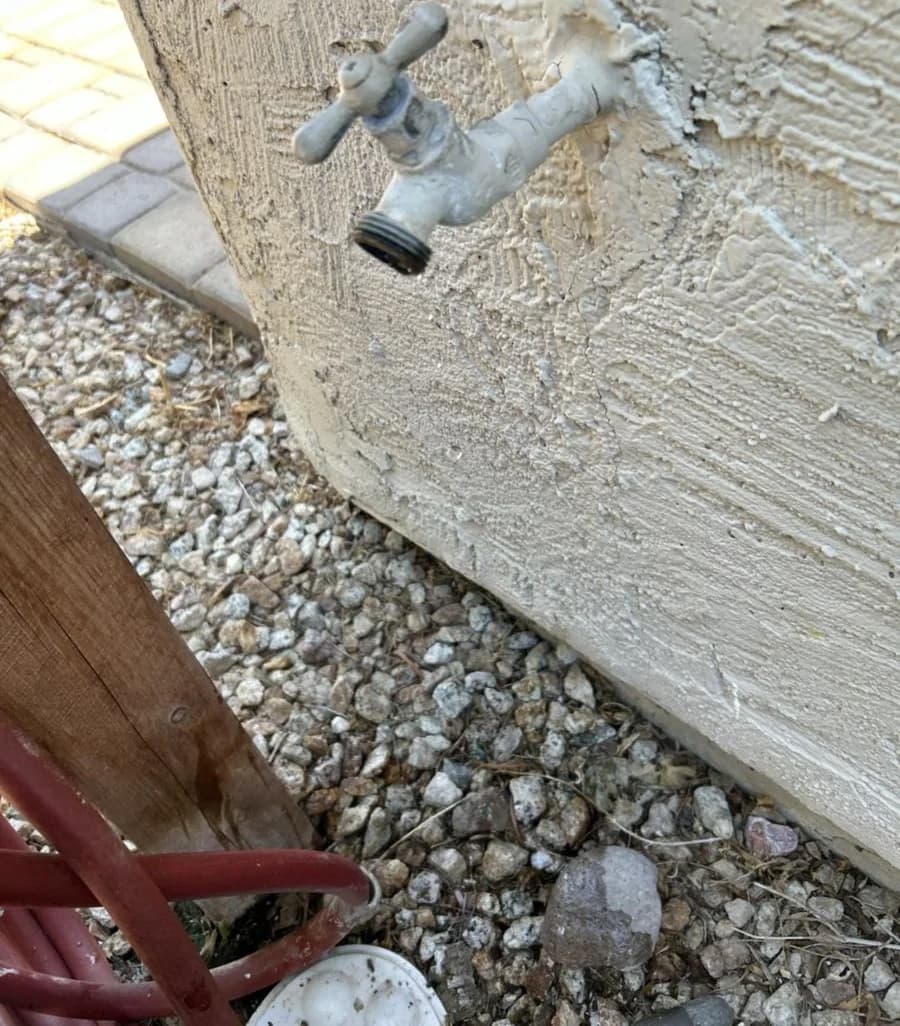 A corroded, paint-crusted chrome hose bib set in patched stucco above desert gravel, before replacement
