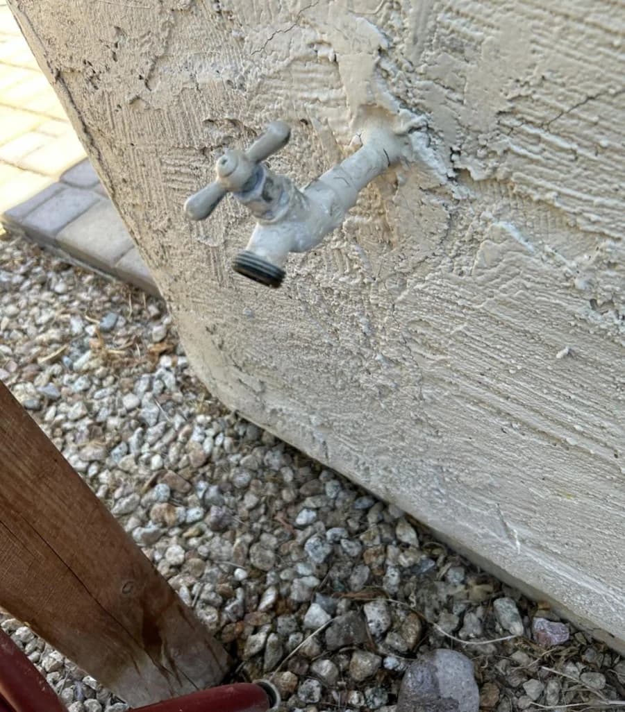 A corroded exterior hose bib crusted into cracked stucco above desert gravel