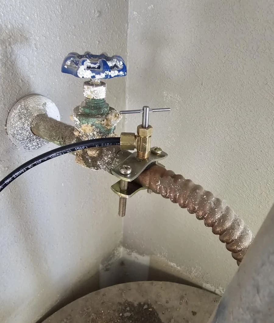 A saddle valve clamped onto a corroded supply line above a water heater, beside a mineral-crusted gate valve