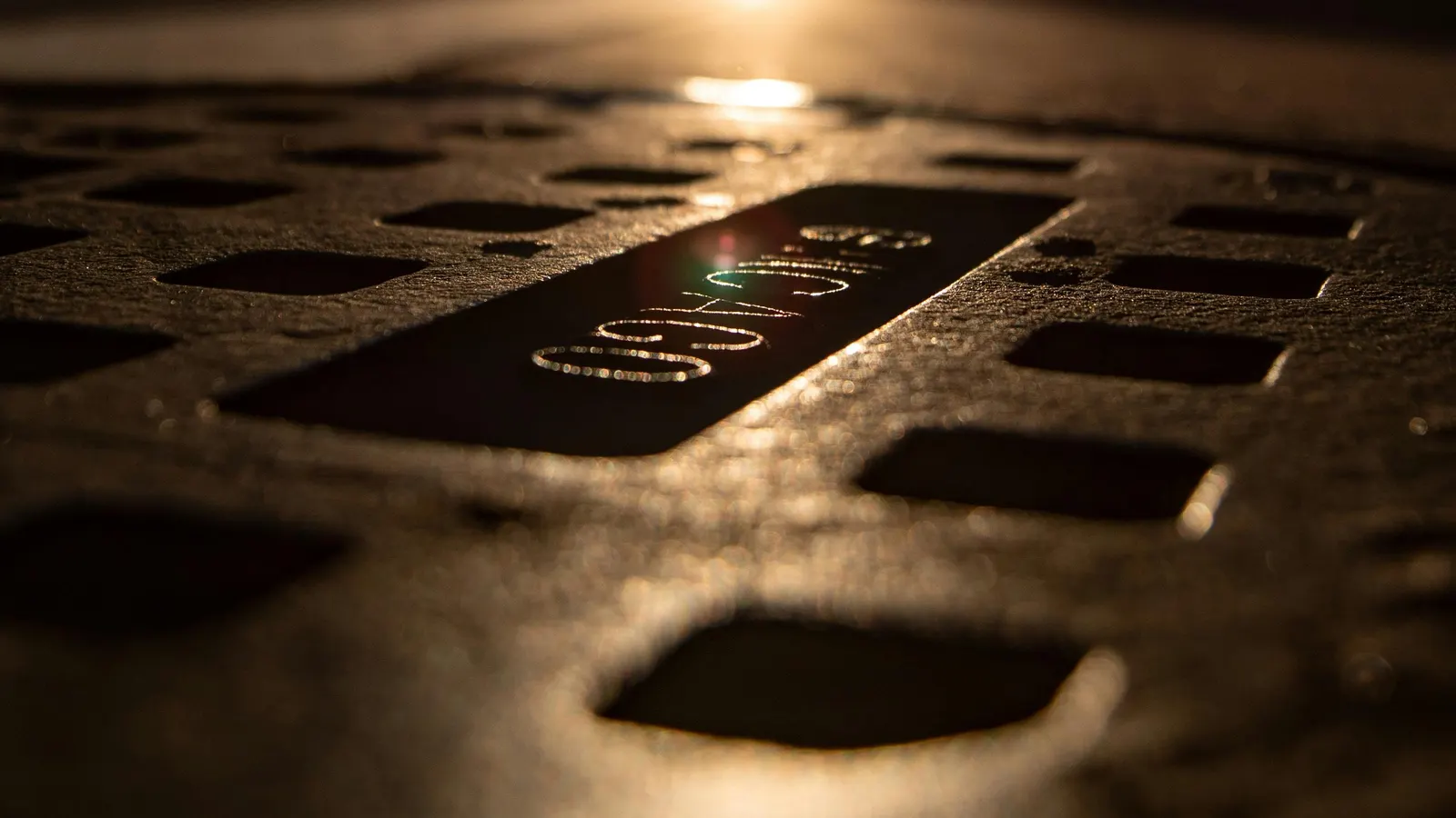 A slotted cast-iron manhole cover with raised lettering, lit by low golden sunlight