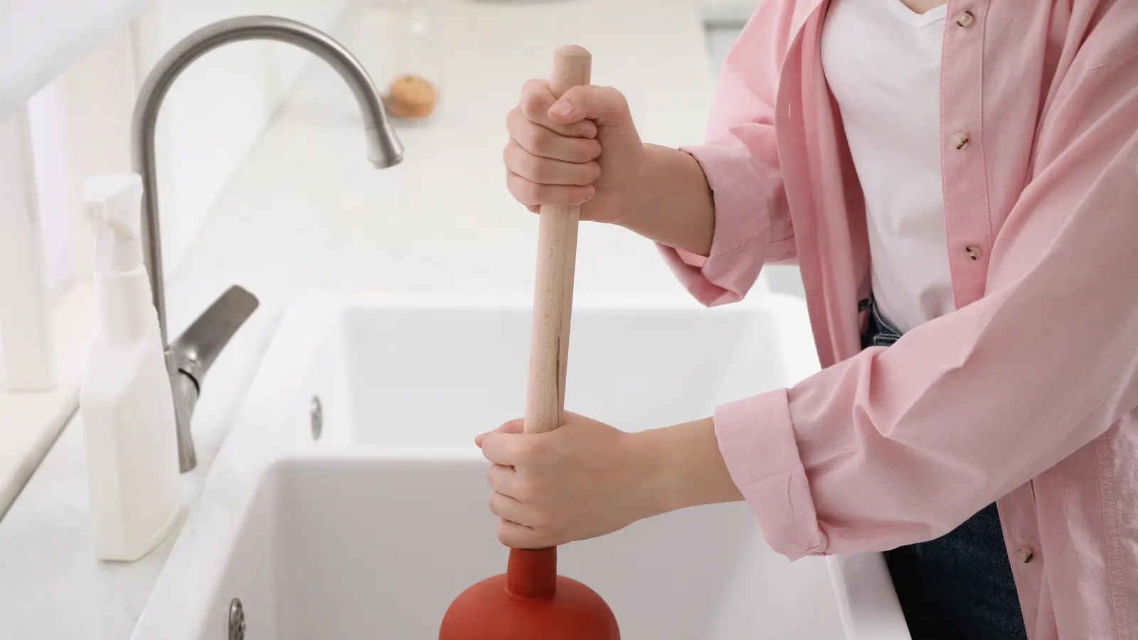 A person in a pink shirt working a red rubber plunger in a white kitchen sink
